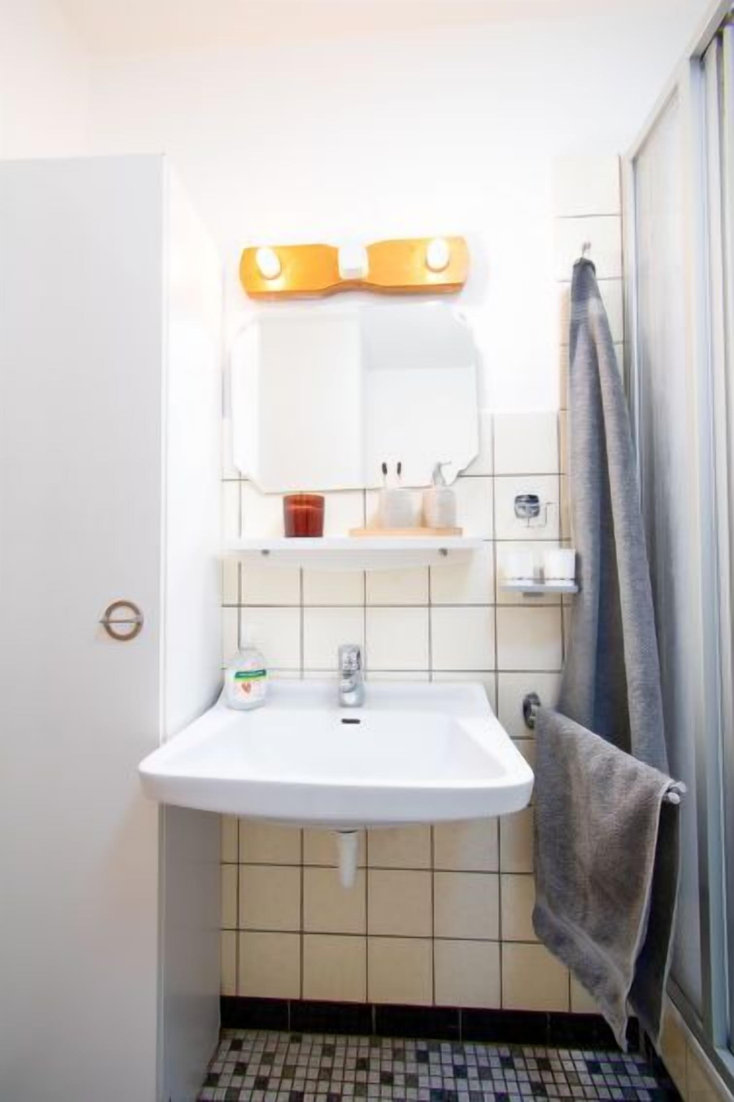 Une salle de bain avec un lavabo blanc, un miroir avec éclairage intégré, un porte-serviettes et un sol carrelé.