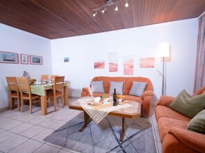 A living room with a wooden ceiling, a dining table with chairs, a sofa, and a floor lamp.