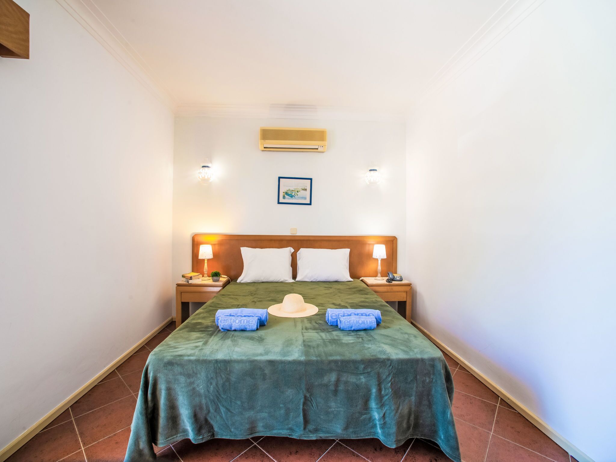Une chambre d’hôtel comprenant un lit, deux tables de chevet, une petite table, un climatiseur fixé au mur et un tableau encadré accroché au mur.