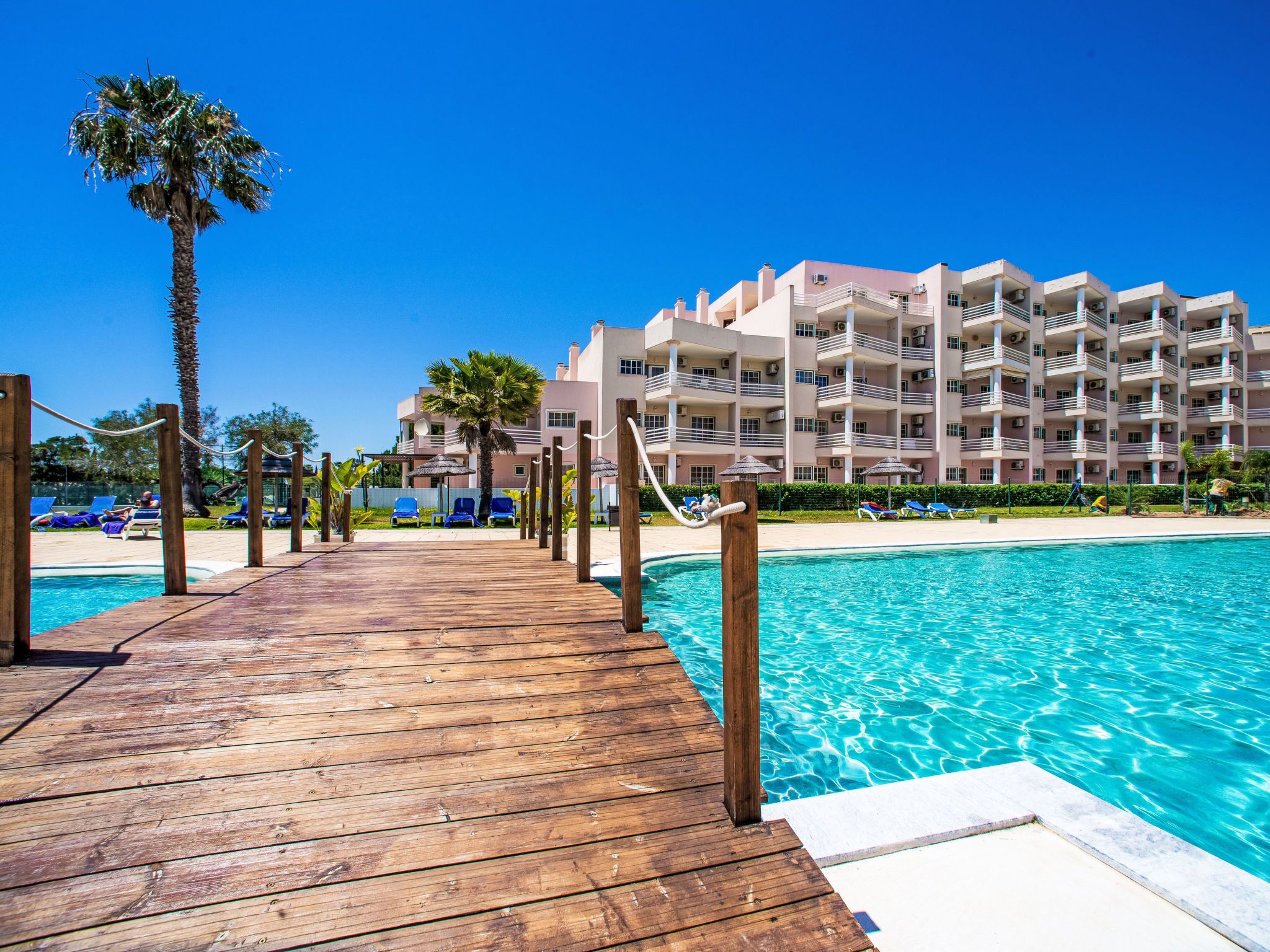 Une terrasse en bois mène à une piscine dont l’eau est d’un bleu cristallin, le tout étant entouré d’un immeuble d’hôtel à plusieurs étages, doté de balcons.