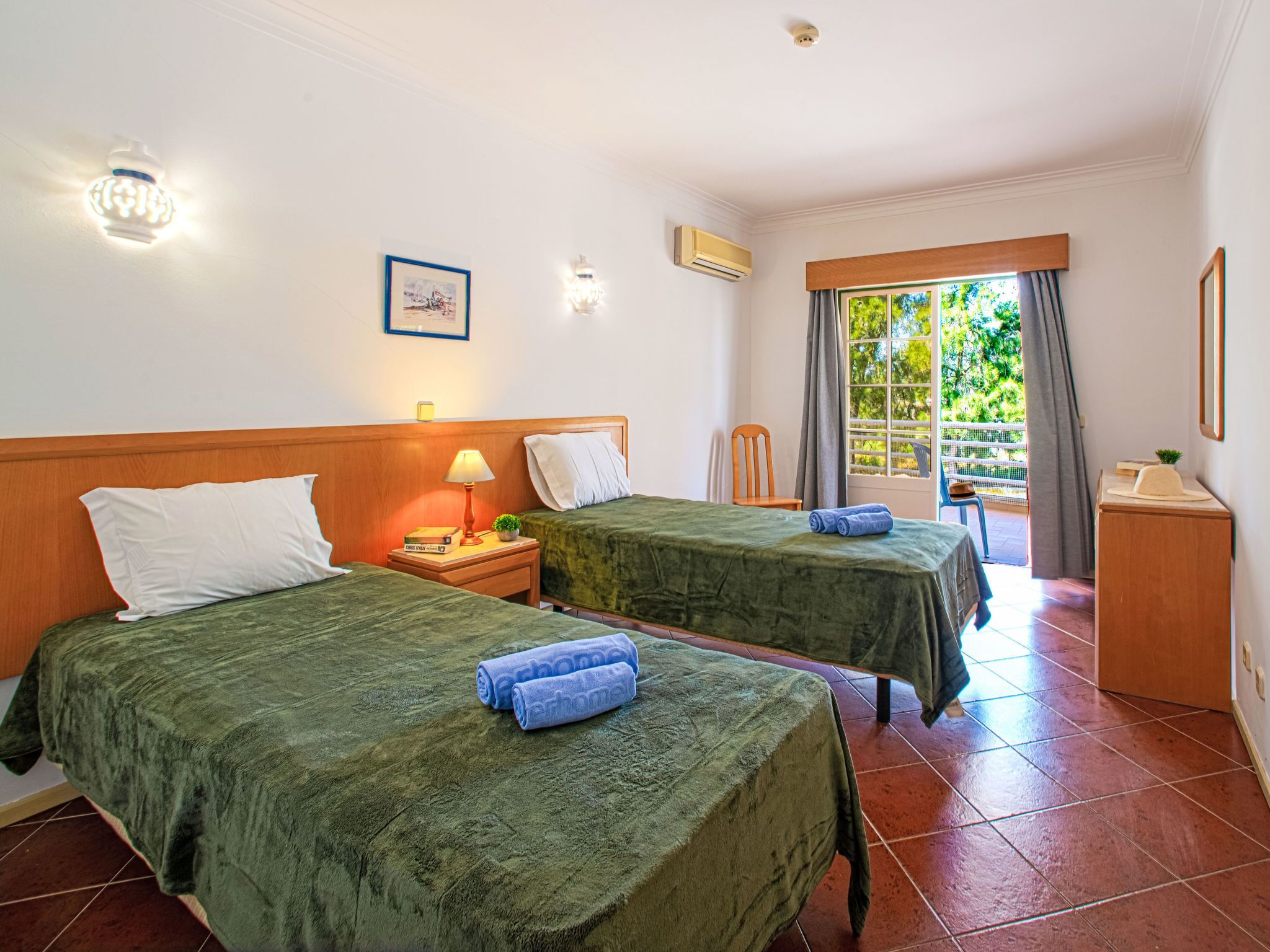 Habitación de hotel con dos camas, una mesita de noche con lámpara, una pequeña mesa con planta y una ventana con cortinas.