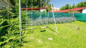 Ein Metall-Schaukelstuhl in einem grasbewachsen Hinterhof mit einem grünen Zaun und Bäumen im Hintergrund.