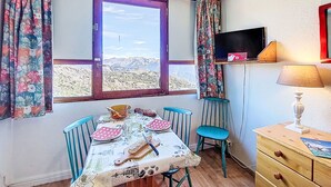 Una habitación con una mesa de comedor dispuesta para dos personas, una ventana con vistas a las montañas, un televisor en una estantería y una lámpara en una cómoda.