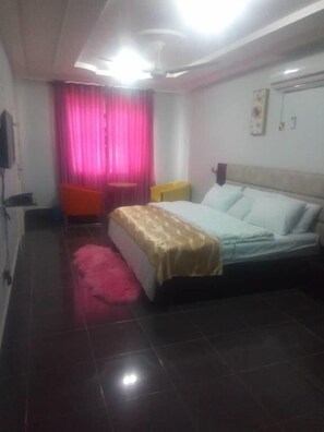 A hotel room with a bed, a desk, a chair, a window with curtains, and a ceiling fan. - Ama Serwaa Lodge (Taifa)