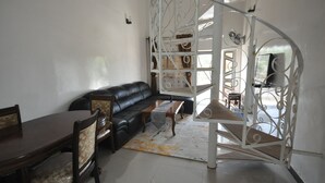 Een woonkamer met een zwarte leren bank, een houten eettafel en stoelen. Er is een witte spiltrap met een metalen leuning.