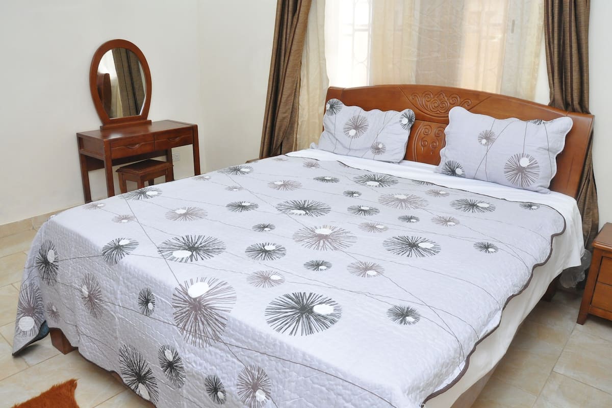 A neatly made bed with a floral-patterned bedspread, a wooden nightstand, a small wooden table with a mirror, and a brown headboard.