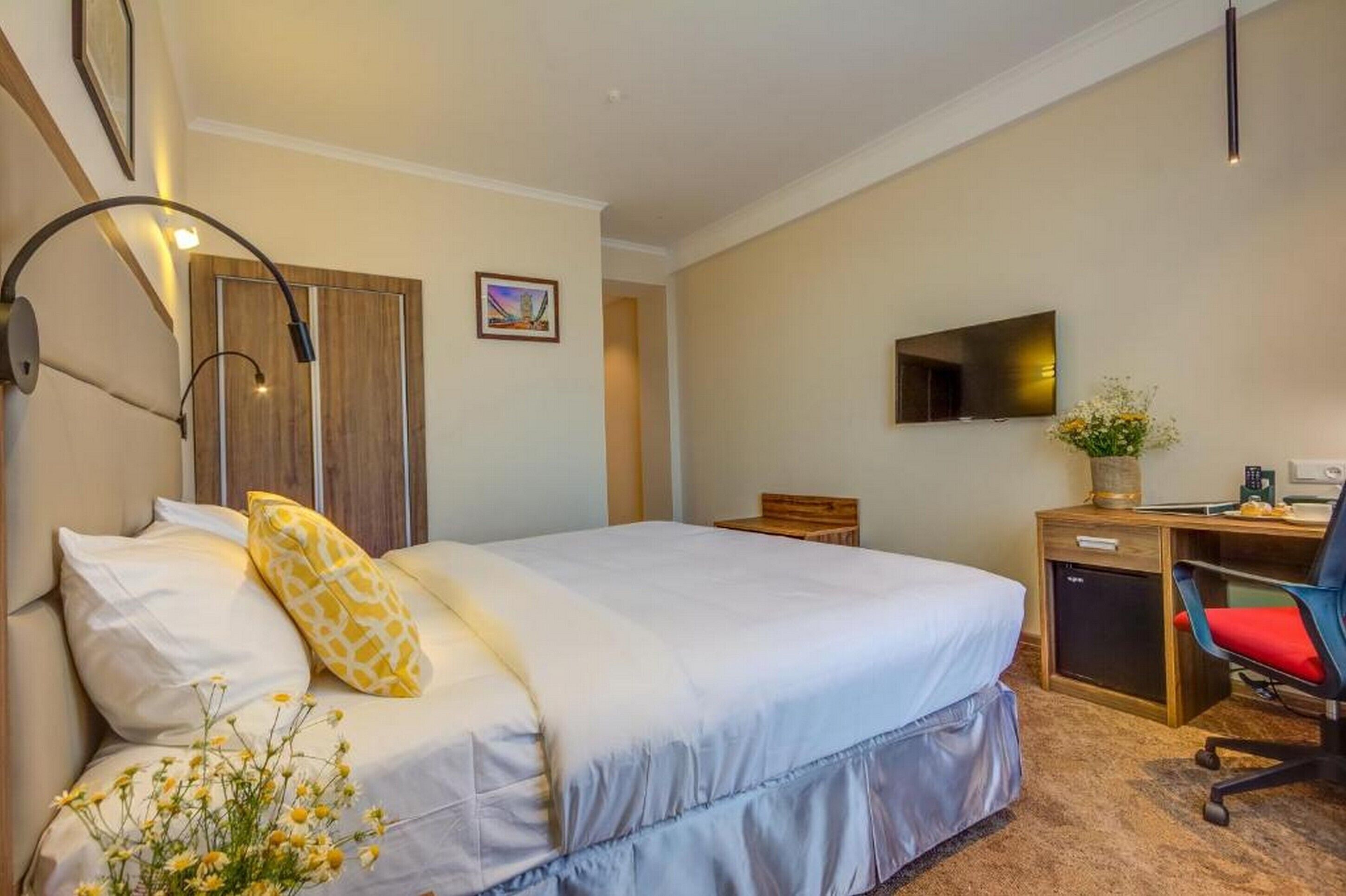 A hotel room with a bed, bedside table, desk, chair, TV, and a small plant.