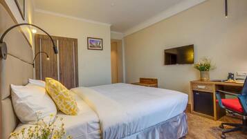 A hotel room with a bed, bedside table, desk, chair, TV, and a small plant.