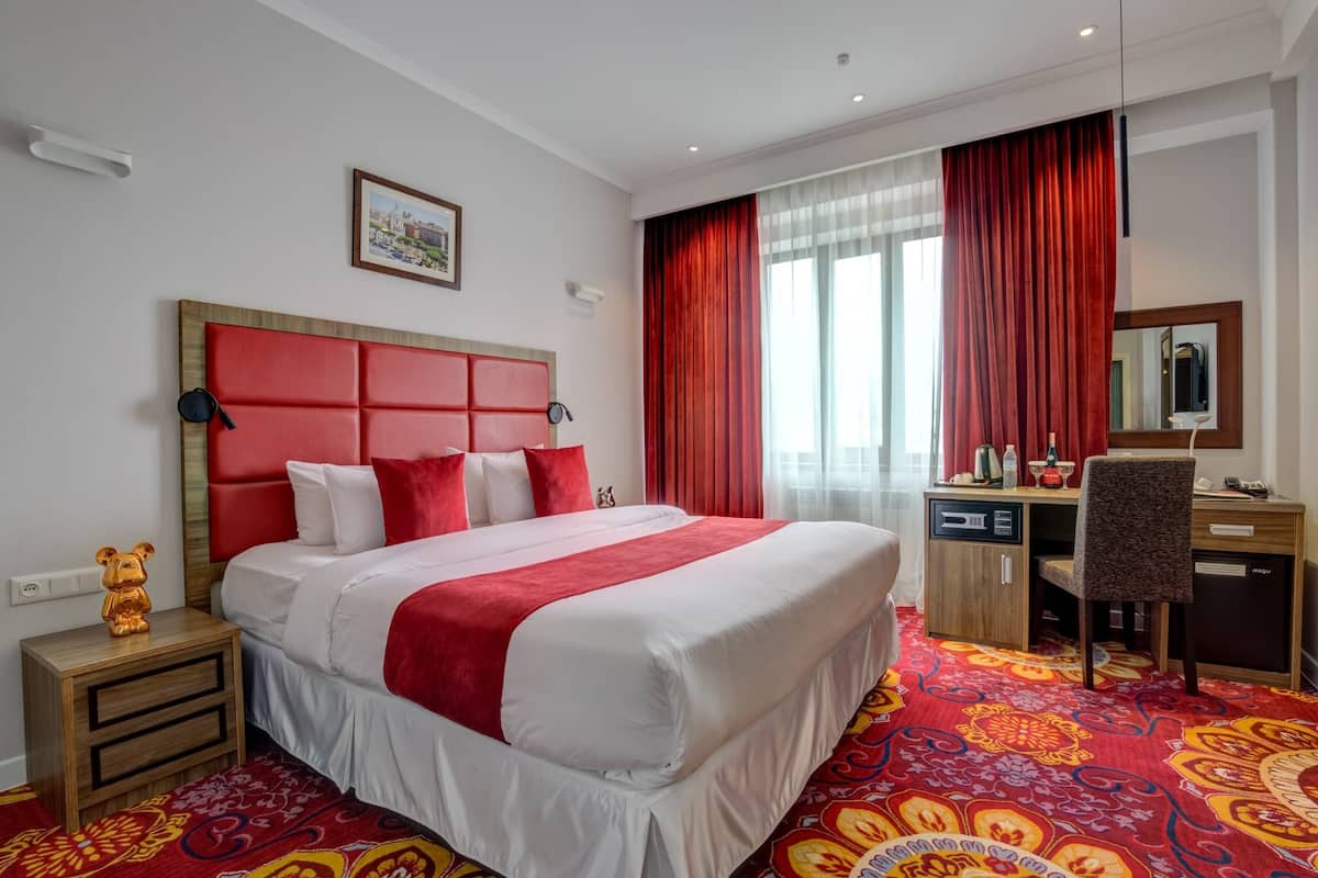 A hotel room with a large bed, a desk, a chair, a nightstand, a red headboard, and a patterned carpet.