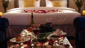 A romantic dinner setup with a bed in the background, candles, and rose petals on the table. - Garden Hotel & Spa (Bishkek)
