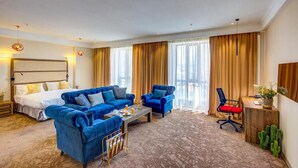 A hotel room with a large bed, two blue sofas, a coffee table, a desk, and a cactus plant. - Garden Hotel & Spa (Bishkek)