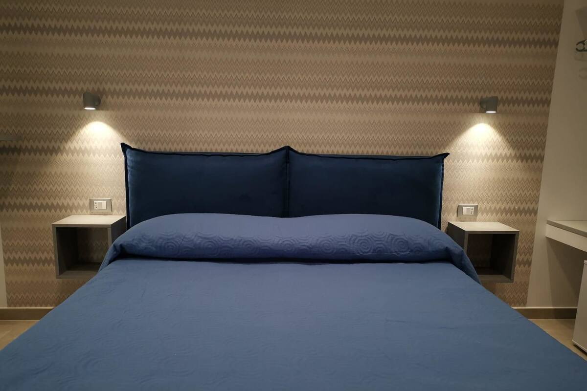 A bed with a blue bedspread and pillows, flanked by two bedside tables with lamps, against a patterned wall.