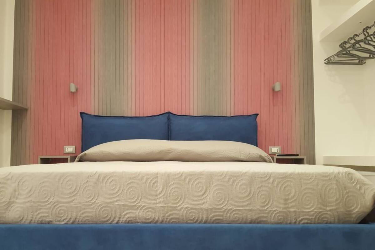 A bed with a beige and blue bedspread, a pink striped wall, and a small shelf with a lamp.