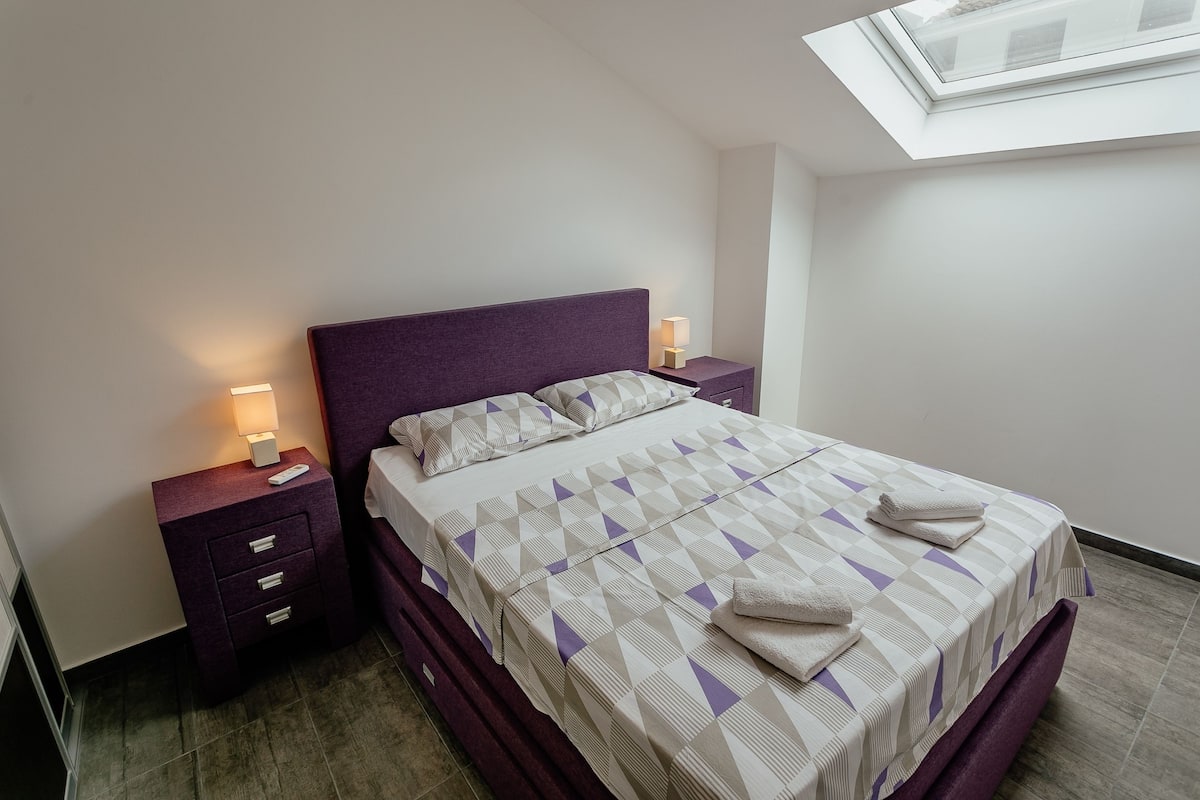 Una cama bien hecha con una colcha de estampado geométrico, un cabecero morado y dos mesitas de noche con lámparas.