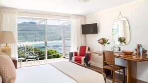 A hotel room with a large bed, a desk, a chair, a TV, and a view of the sea and mountains. - Poseidon Guest House (Cape Town)