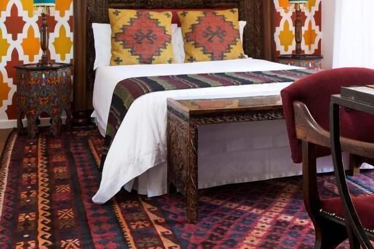 Un dormitorio con pared estampada, una cama de madera, dos mesitas de noche con lámparas, una silla roja y una alfombra con diseño.