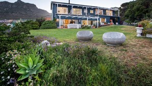 A modern two-story house with a covered patio, surrounded by a well-maintained garden with various plants and a large rock feature. - Poseidon Guest House (Cape Town)
