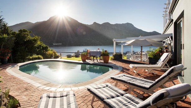 A swimming pool with lounge chairs, a table, and a view of mountains and the sea. - Poseidon Guest House (Cape Town)