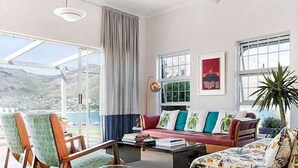 A spacious living room with a large window offering a view of the outdoors, a red sofa with patterned cushions, and a wooden coffee table. - Poseidon Guest House (Cape Town)