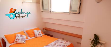 Un dormitorio con cama, almohadas, mesita de noche, una silla y una ventana.