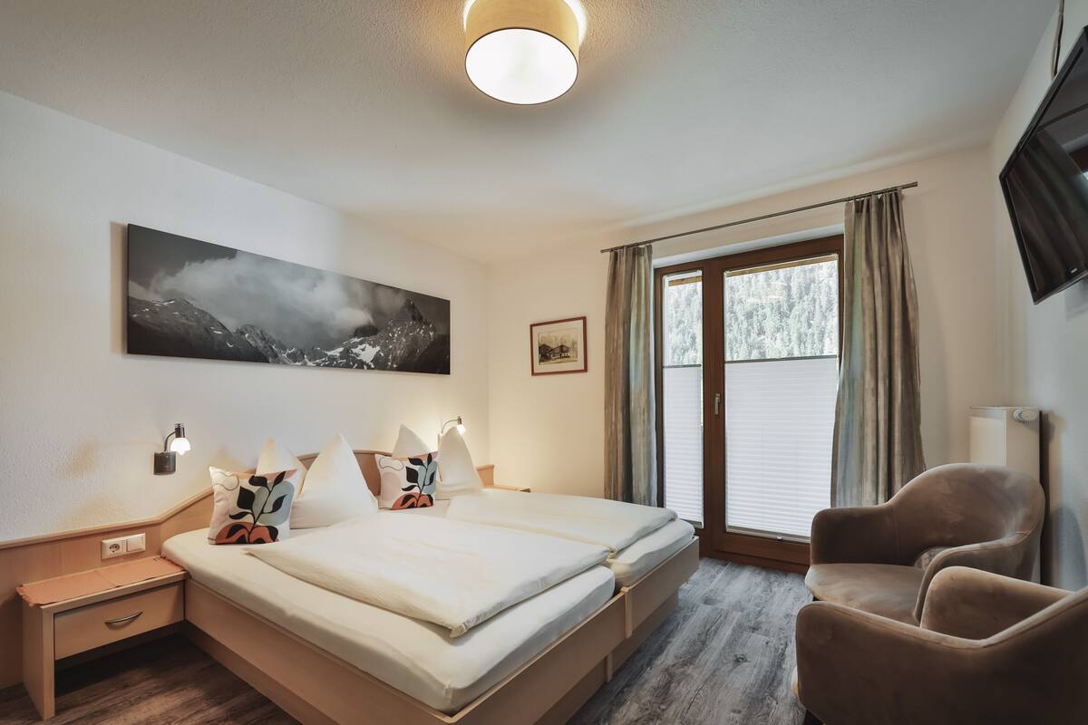A hotel room with a bed, bedside tables, a chair, a large window with curtains, a painting on the wall, and a television.
