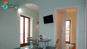 A dining area with a table and chairs, a flat-screen TV mounted on the wall, and a view of the outside through a door. - Casa Ares Sperlongaresort (Sperlonga)