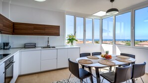 Eine moderne Küche mit weißen Schränken, einem Esstisch mit Stühlen und großen Fenstern mit Blick auf den Ozean.