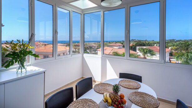 Ein Essbereich mit Tisch für vier Personen, große Fenster mit Blick auf das Meer und Gebäude, und eine Vase mit Blumen auf der Fensterbank.