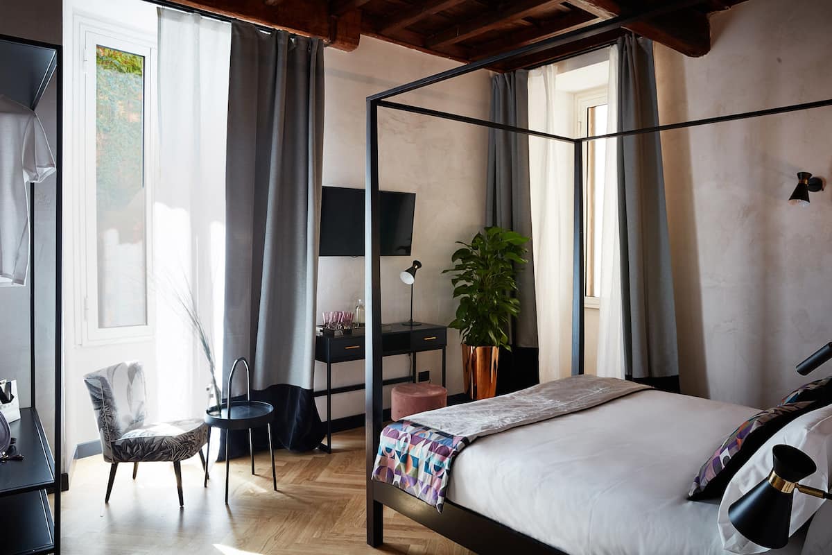 A four-poster bed with a white bedspread, a desk with a computer and a plant, a chair, and a television on the wall.