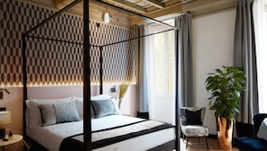 A four-poster bed with a geometric patterned headboard, a bedside table with a lamp, a potted plant, and a chair with a patterned cushion. - Naman Hotellerie Via Margutta (Rome)