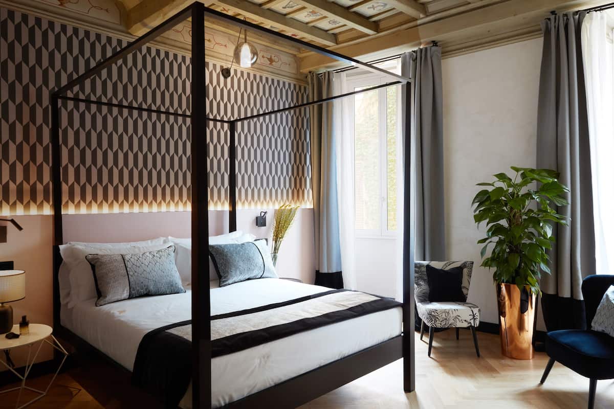 A four-poster bed with a geometric patterned headboard, a bedside table with a lamp, a potted plant, and a chair with a patterned cushion.