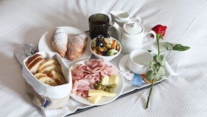A breakfast tray with croissants, bread, ham, cheese, olives, and a fruit salad on a bed. - Naman Hotellerie Via Margutta (Rome)