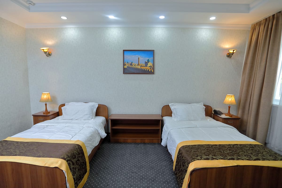 A hotel room with two beds, a nightstand with a lamp, a wall-mounted picture, and a window with curtains.