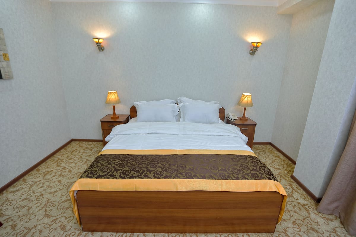 A neatly made bed with white linens and a brown headboard, flanked by two bedside tables with lamps, in a room with patterned carpet and light-colored walls.