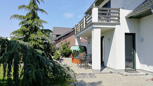 A modern building with a balcony, a paved courtyard, and a large coniferous tree. - Appartements Süssling (Glowe)