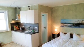 A compact hotel room with a kitchenette, a bed with white bedding, a nightstand with a lamp, and a painting on the wall. - Appartements Süssling (Glowe)