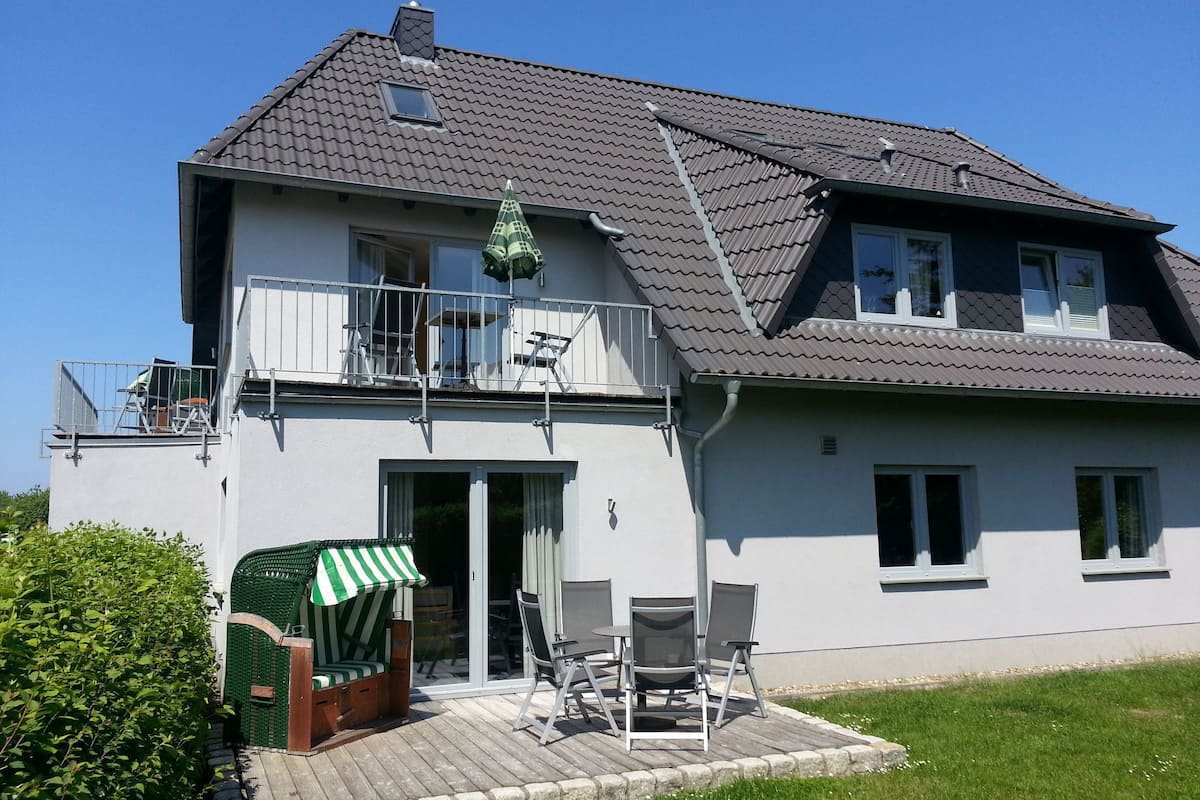 Une maison à deux étages avec un balcon, une terrasse couverte aménagée avec du mobilier d’extérieur et un portail vert.