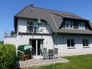 Une maison à deux étages avec un balcon, une terrasse couverte aménagée avec du mobilier d’extérieur et un portail vert.