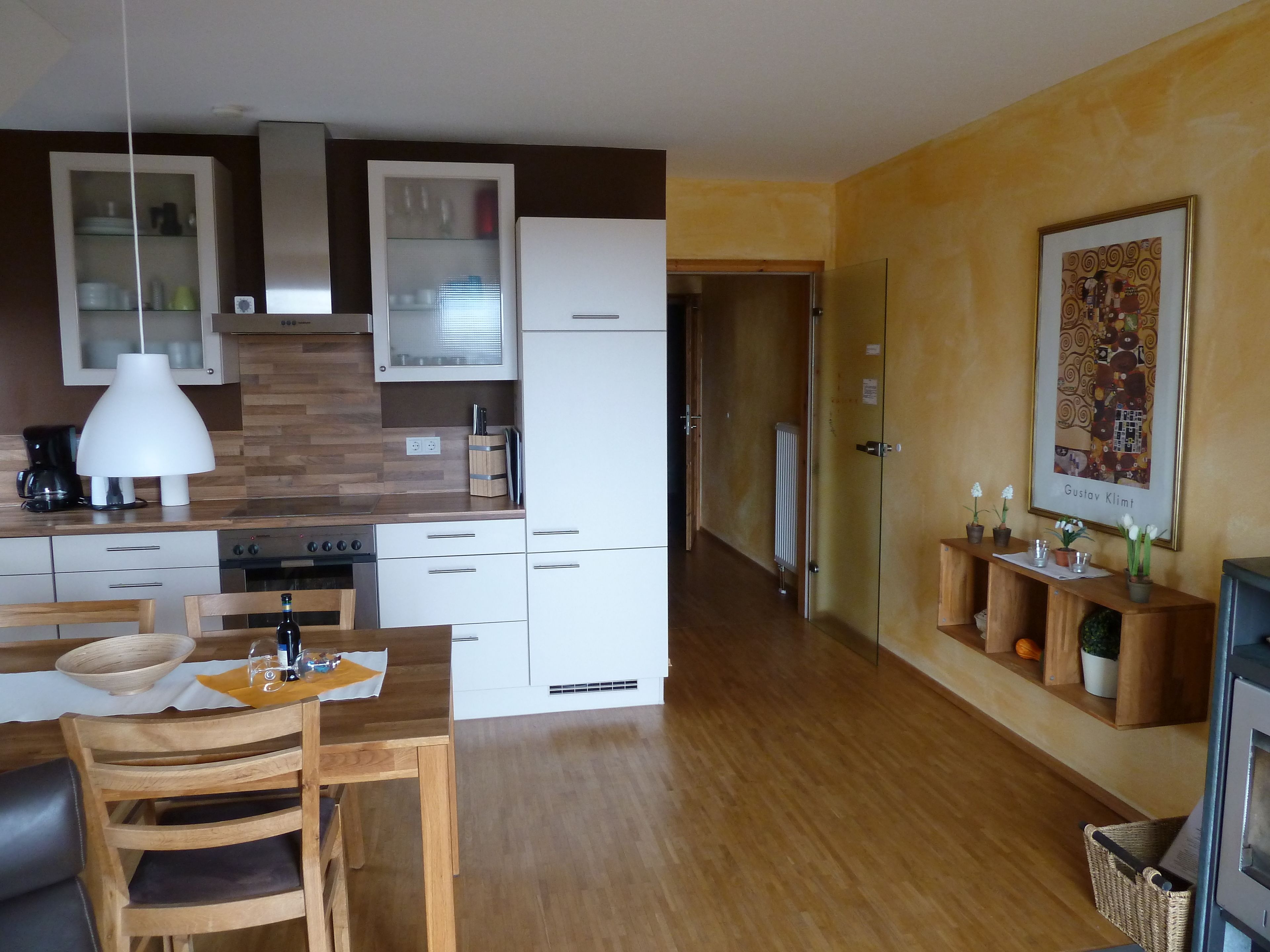 Una cocina moderna con armarios blancos, encimera de madera y una lámpara colgante. Hay un comedor con una mesa y sillas de madera.