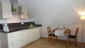 Eine kompakte Küche mit Kaffeemaschine, Spüle und einer kleinen Pflanze auf der Arbeitsfläche. Im Eck befindet sich ein Esstisch mit zwei Stühlen und einer Lampe.