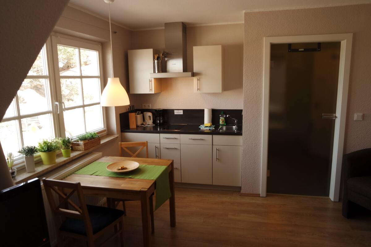 A compact kitchen with a dining area, a window with plants, and a door leading to another room.