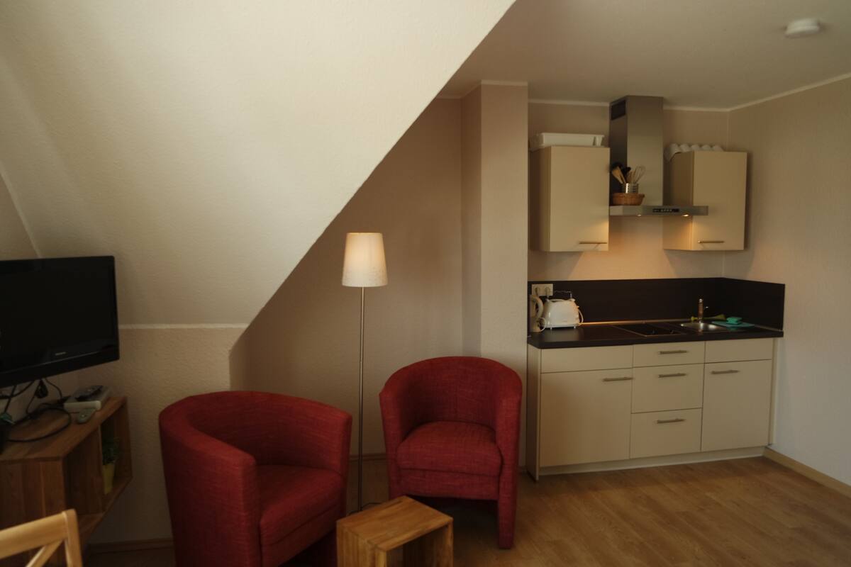 A compact living space with a kitchenette, two red armchairs, a small wooden table, and a television.