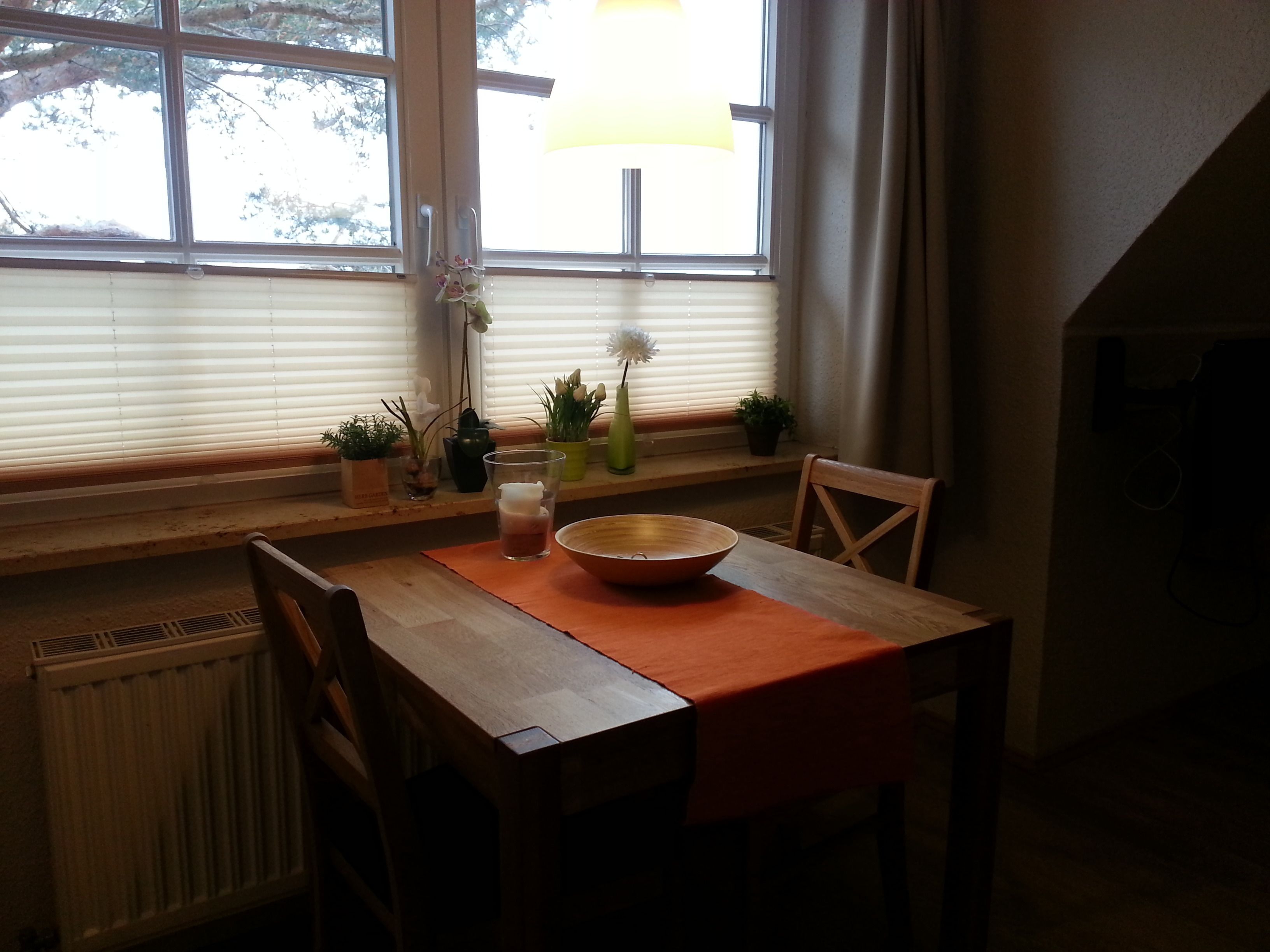 Een eetgedeelte met een tafel, stoelen, een schaal, een vaas met bloemen en potplanten op een vensterbank.