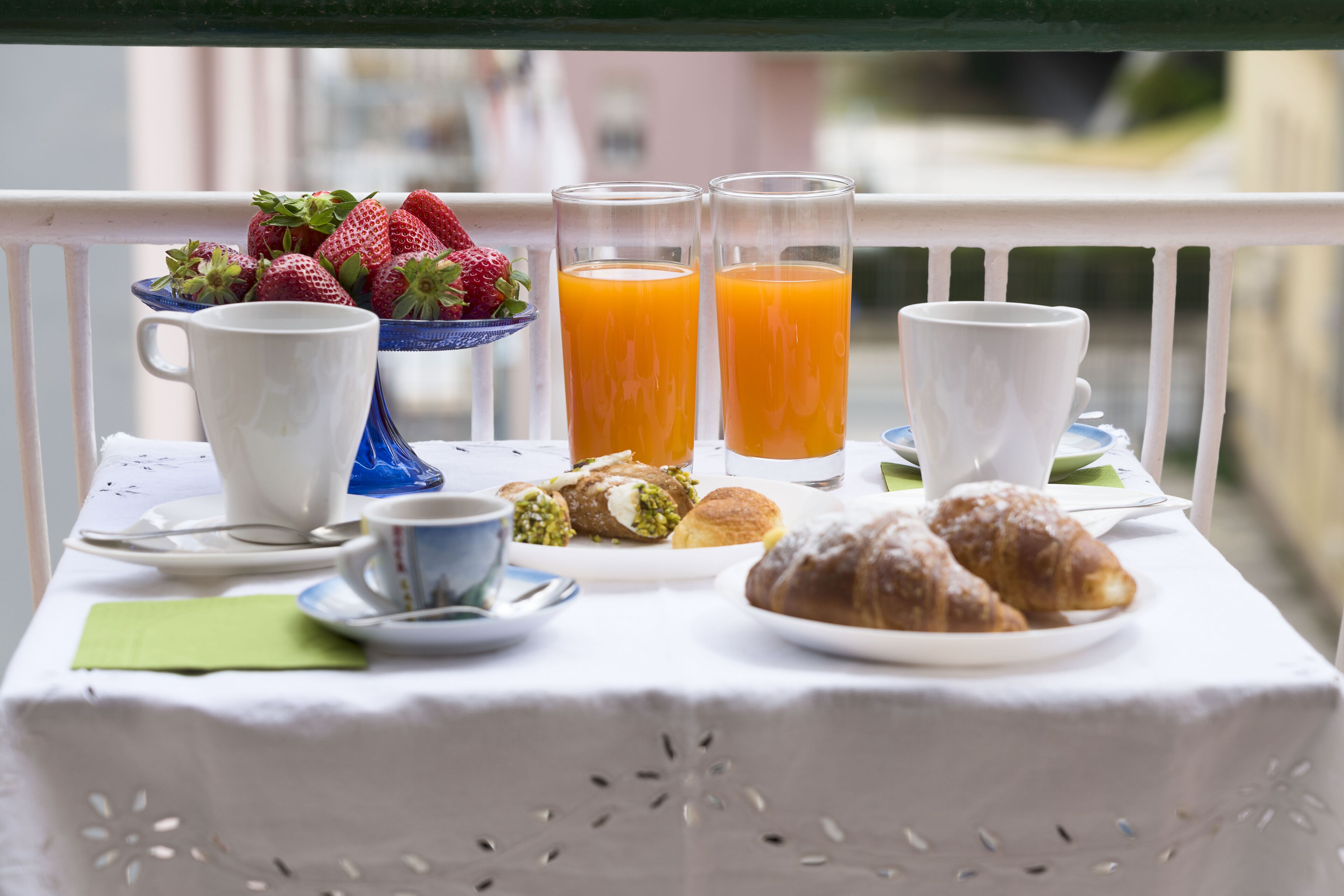 A breakfast table with a white tablecloth, a bowl of strawberries, two glasses of orange juice, a cup of coffee, a plate with pastries, and a green napkin.