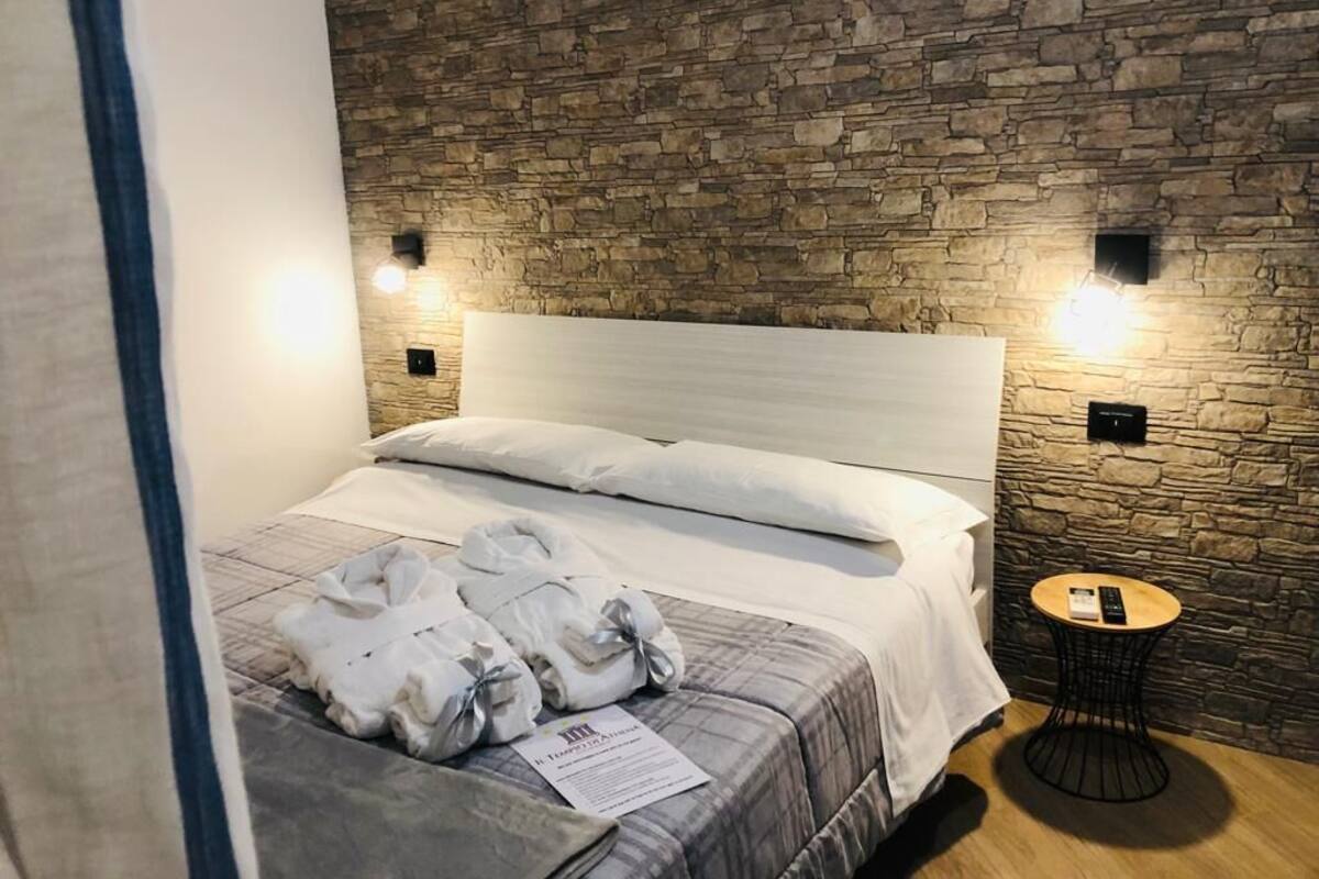 A hotel room with a stone wall, a bed with white bedding, a small round table with a remote control, and a note on the bed.