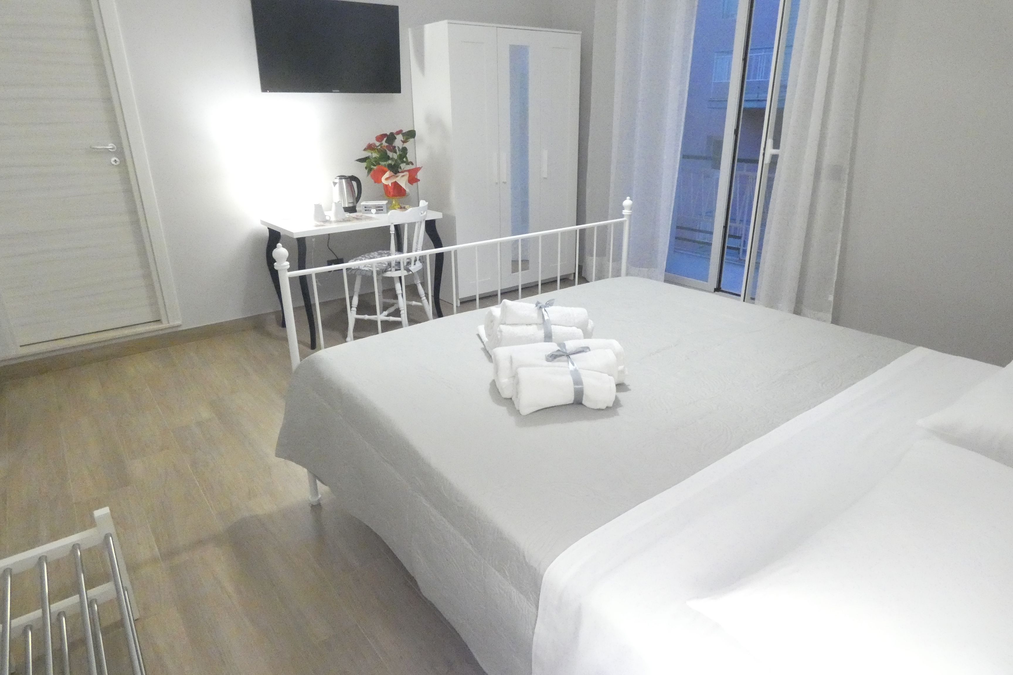 A hotel room with a neatly made bed, a desk with a plant, a wardrobe, and a view of the outside through a window.