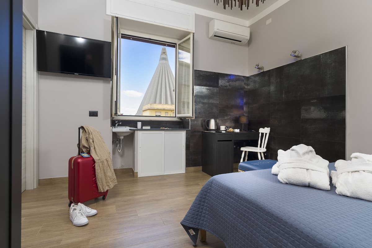 A modern hotel room with a bed, a desk, a TV, a window with a view of a building, and a red suitcase.