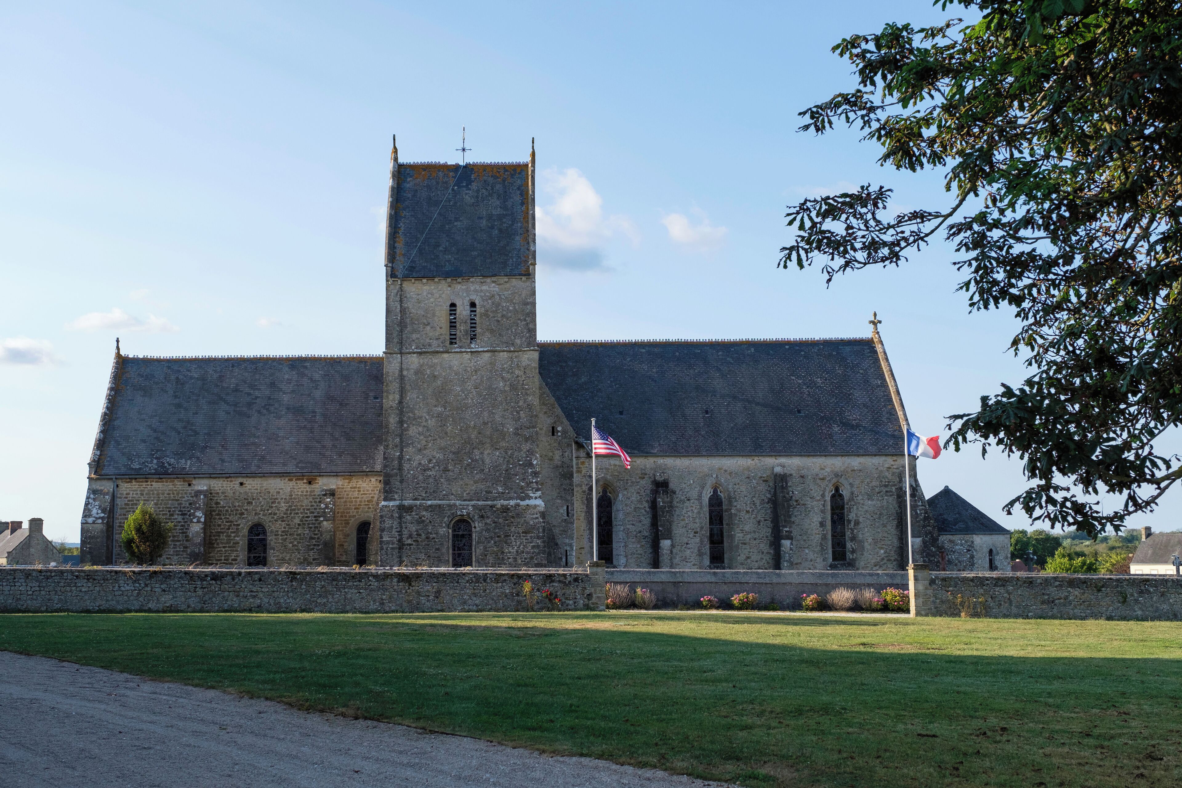 A historic stone building with a prominent tower, a flagpole with two flags, and a well-maintained lawn.