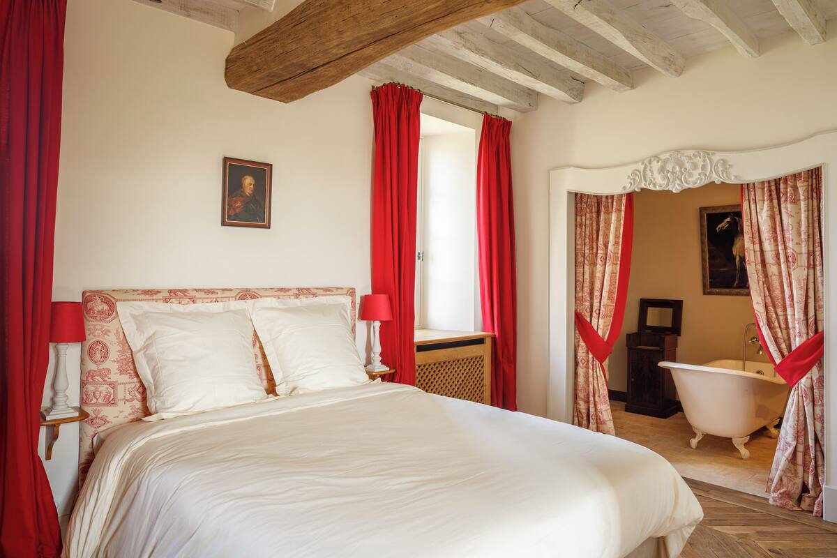 Una camera da letto con un letto, tende rosse, una testiera in legno, un quadro incorniciato al muro e un bagno con una vasca e uno specchio.