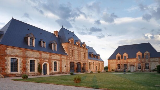 A large, historic stone building with multiple gabled roofs and arched windows, surrounded by a well-maintained lawn. - Le Domaine de Ravenoville (Sainte-Mere-Eglise)